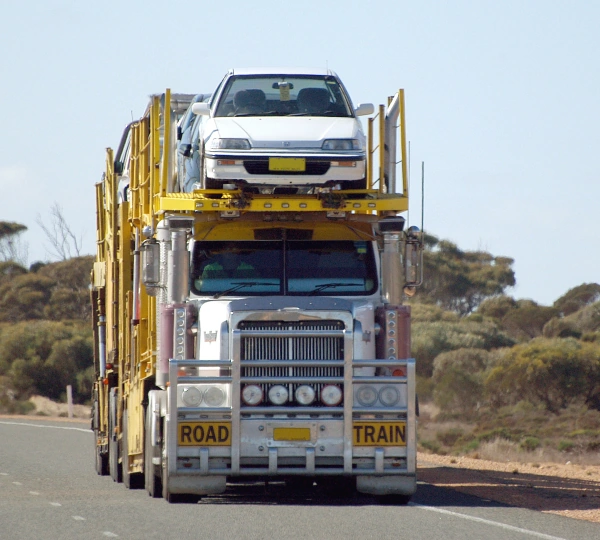Large road train truck transporting multiple vehicles, with a white car on top, on a rural road with sparse vegetation.